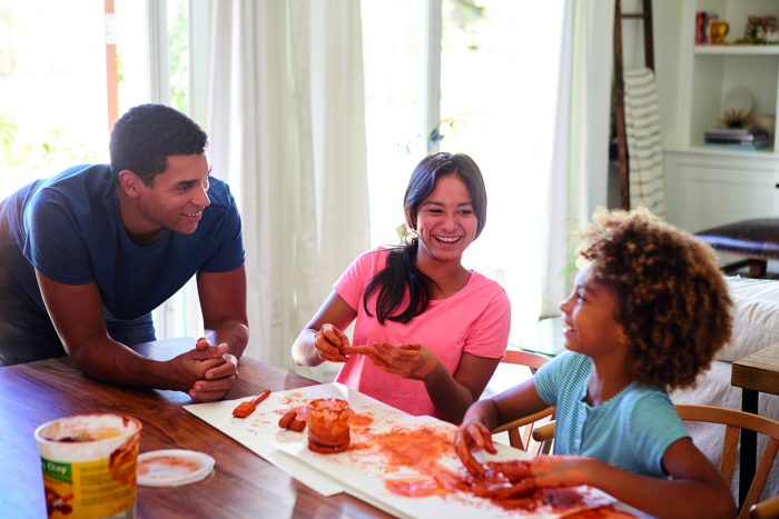 Millennial dad leaning on table watching his daughter and her gi Image of a father leaning on a table with two girls one older than the other playing with modelling clay.
