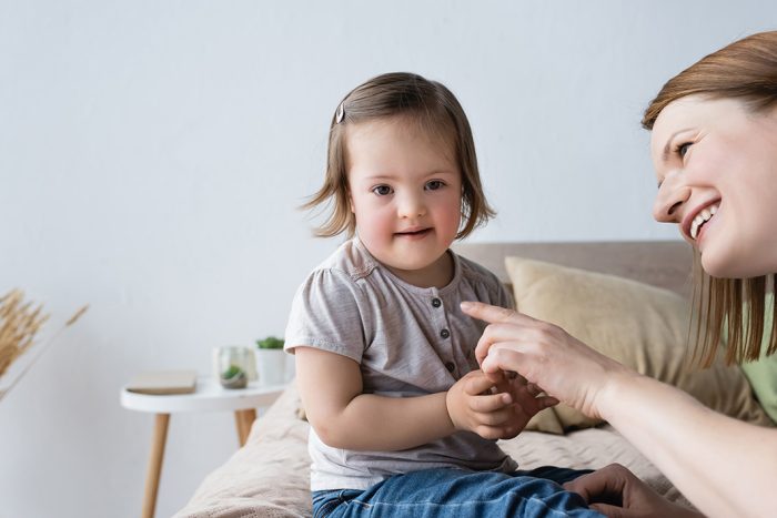 Smiling Mother Pointing With Finger At Toddler Daughter With Dow Smiling mother pointing with finger at toddler daughter with down syndrome in bedroom