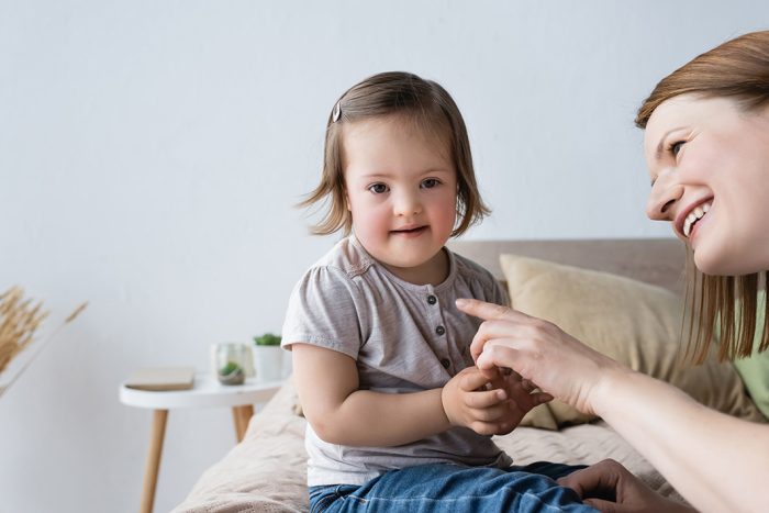 Smiling mother pointing with finger at toddler daughter with down syndrome in bedroom